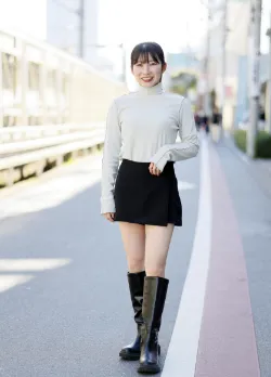 グレーのトップスと黒いミニスカート、ロングブーツを着用し、街中でポーズを取る女性ファッションモデル